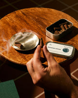A hand holds a rolled incense cone over a smoking metal dish on a round wooden table, with an open tin of incense cones and a branded product box nearby.