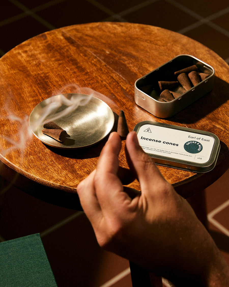 A hand holds a rolled incense cone over a smoking metal dish on a round wooden table, with an open tin of incense cones and a branded product box nearby.