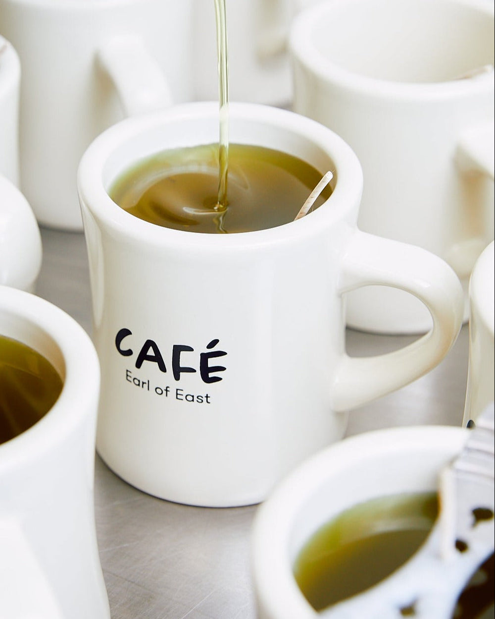 Iced Matcha Scented Candle in Mug, 300ml by Earl of East, lit, on a café table with coffee cups