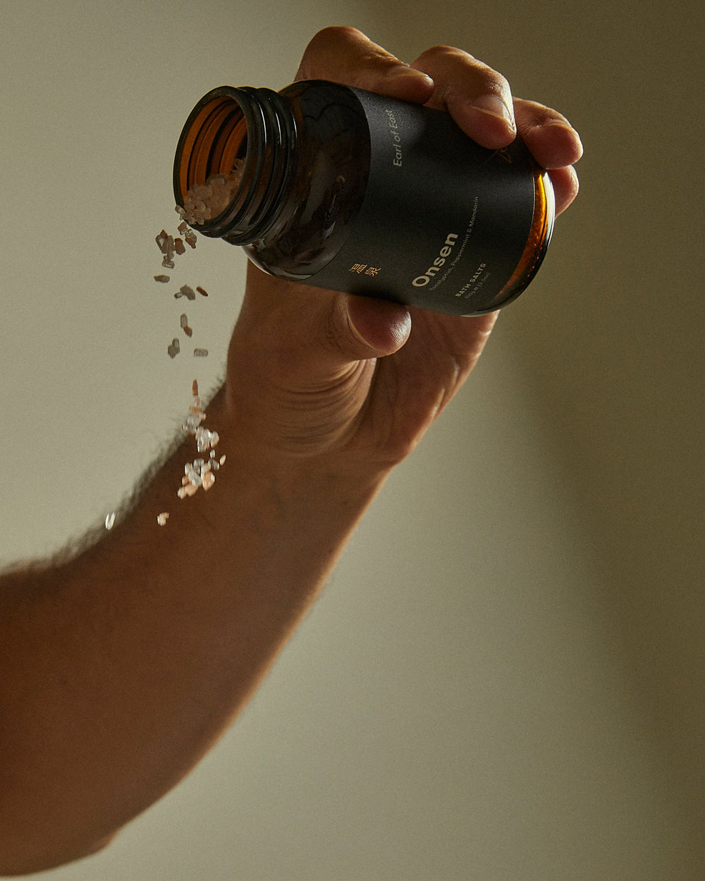 A hand holding an amber glass bottle labeled 'Oraen' with capsules spilling out against a warm beige background.