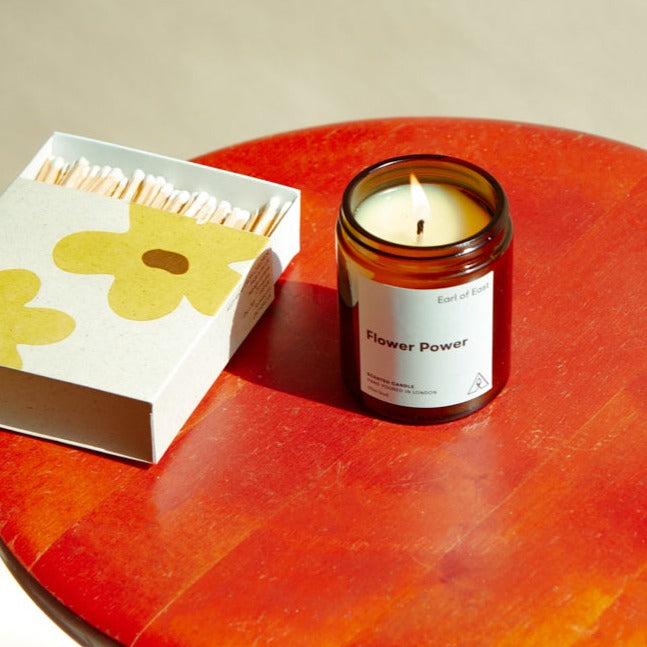 Flower Power, Soy Wax Candle, 170ml [6oz] by Earl of East, lit beside its floral box on a red table