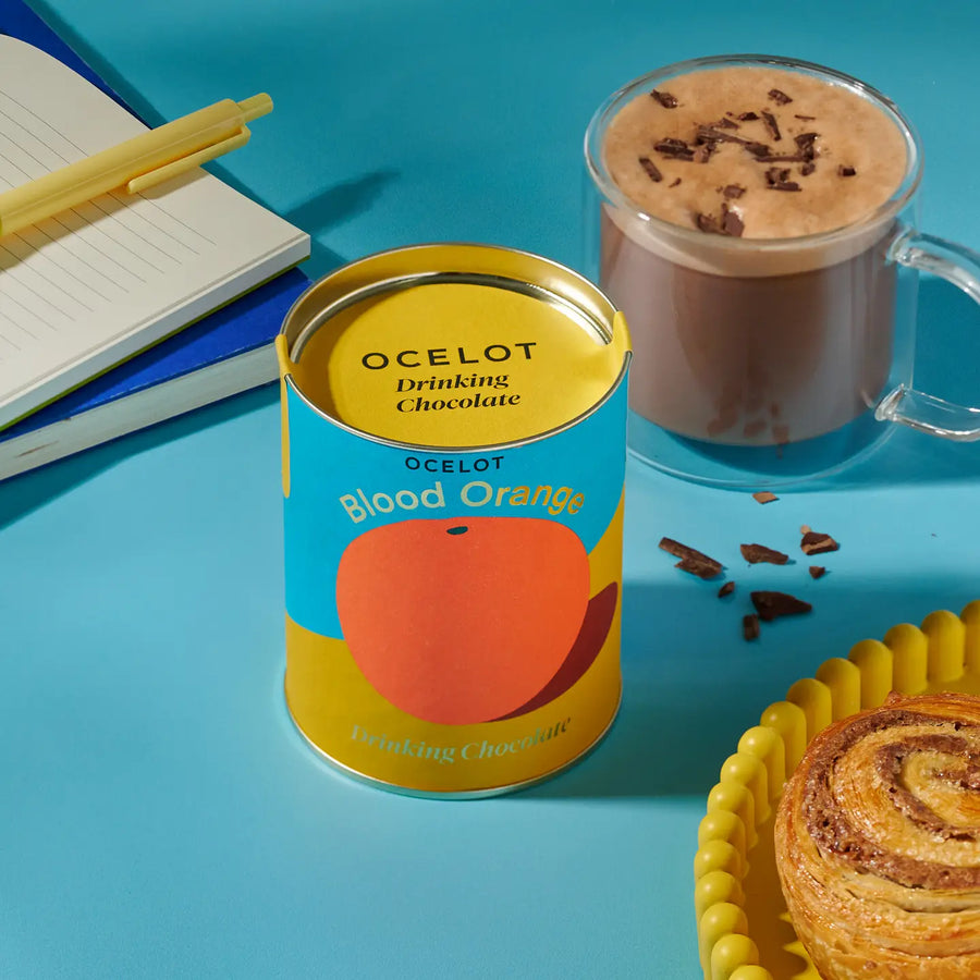 Blood Orange Drinking Choc, Organic Dark Choc Flakes, 200g by Ocelot Chocolate, with a mug of hot chocolate on blue